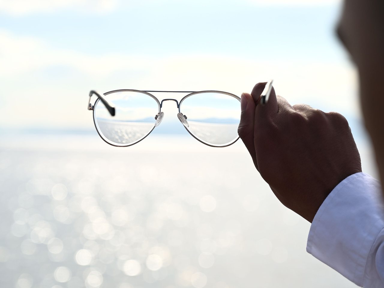 Una mano sostiene unos lentes frente a un horizonte luminoso; el paisaje se ve nítido a través de los cristales